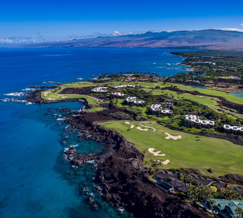 An aerial view of a coastal golf course with lush green fairways and surrounding luxury homes, set against a backdrop of blue ocean and mountains.