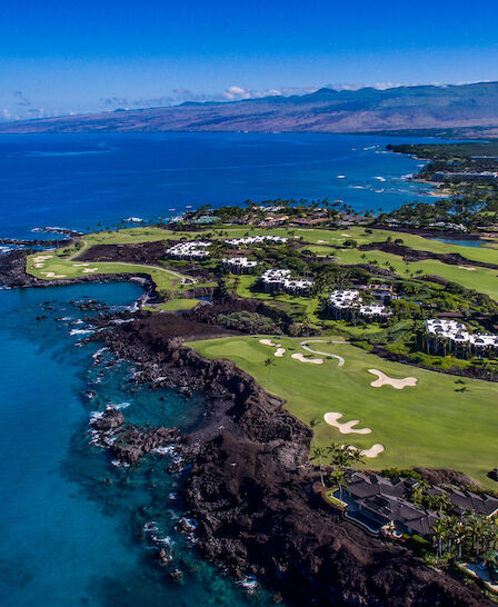 An aerial view of a coastal golf course with lush green fairways and surrounding luxury homes, set against a backdrop of blue ocean and mountains.