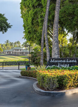 The image shows an entrance to a gated community called "Mauna Lani Point," surrounded by lush greenery and palm trees, with buildings in the distance.