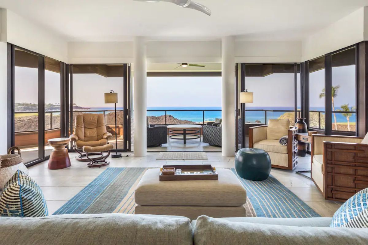 A modern living room with a sea view, featuring cozy furniture, rugs, and wooden decor, leading to a balcony with outdoor seating and ocean views.