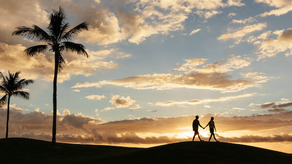 Two people holding hands and walking on a grassy hill during sunset, with palm trees and a colorful sky with clouds in the background.