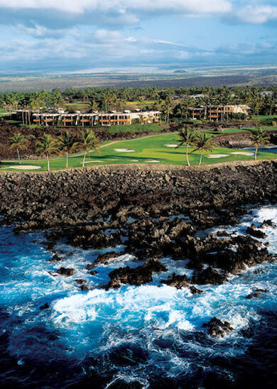 The image shows a coastal golf resort with waves crashing against rocks in the foreground and green fairways, buildings, and palm trees in the background.