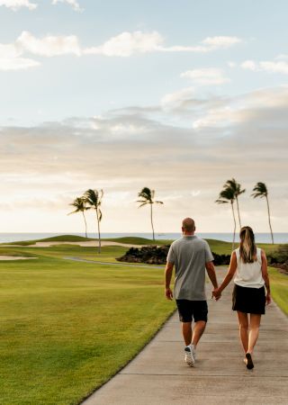 A couple holding hands walks down a path through a scenic, grassy landscape with palm trees and an ocean view in the background.