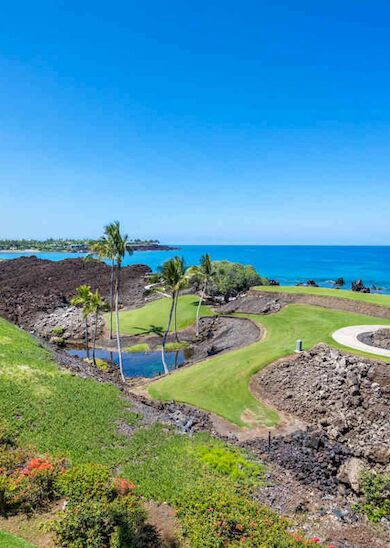 A coastal golf course features a winding path, lush green grass, palm trees, and a stunning ocean view under a clear blue sky.