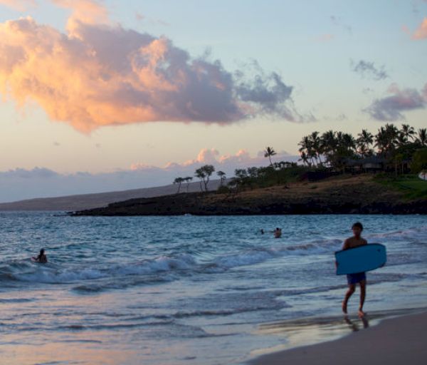 A serene beach scene at sunset with silhouetted palm trees, swimmers in the ocean, and a person carrying a surfboard walking along the shore.