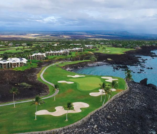 Aerial view of a coastal golf course with sand bunkers, green fairways, buildings, and a rocky shoreline leading to the blue ocean under a cloudy sky.