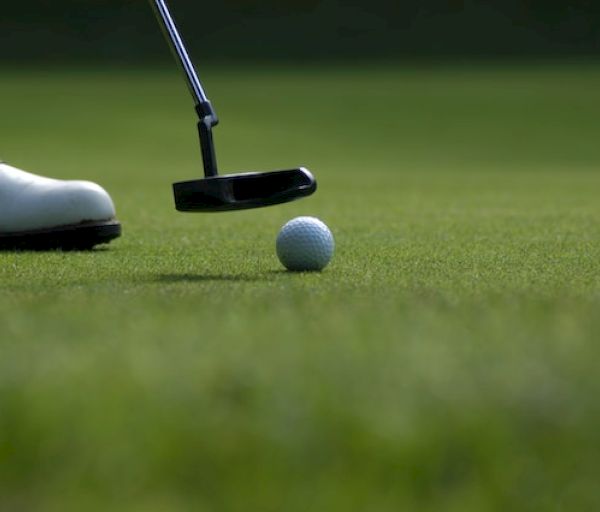 A close-up of a golf ball on a putting green, with a golf club preparing to hit it and a golfer's shoe visible on the left side of the image.