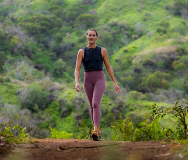 A person is walking on a trail surrounded by lush greenery and hills, wearing athletic clothing and sneakers, looking relaxed and focused.