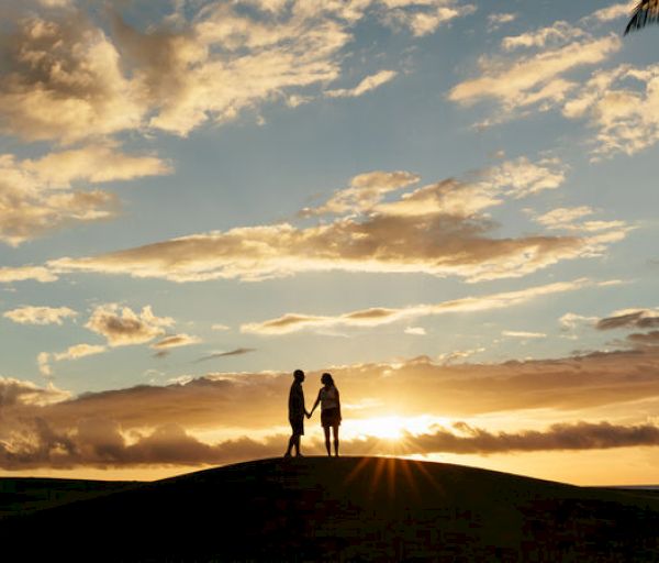 Two people hold hands on a hill during a sunset, with a beautiful sky filled with clouds.