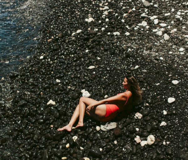 A person in a red outfit is sitting on a rocky beach near the water, surrounded by dark stones and a few white rocks.