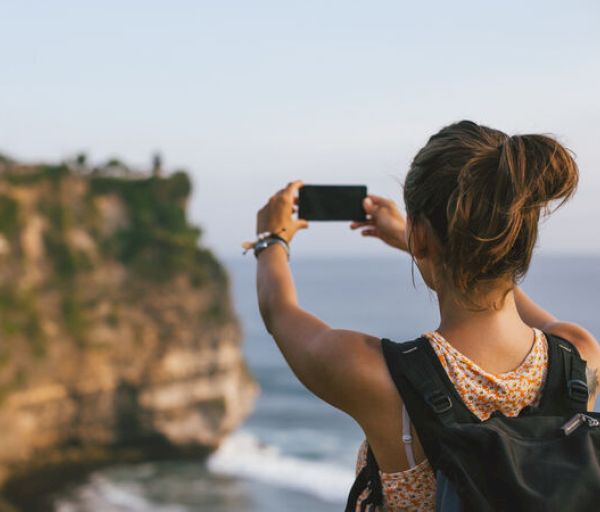 A person with a backpack is taking a photo of a scenic cliff and ocean view using a smartphone, enjoying the beautiful natural landscape.