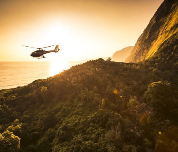 A helicopter flies over lush green hills and a coastline at sunset, with a dramatic cliff to the right and a glowing sky in the background.