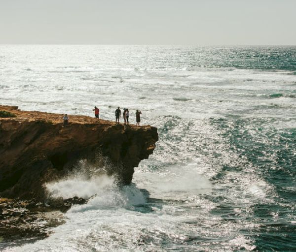 Five people stand on a cliff's edge overlooking a vast, rough ocean with waves crashing against the rocks below.