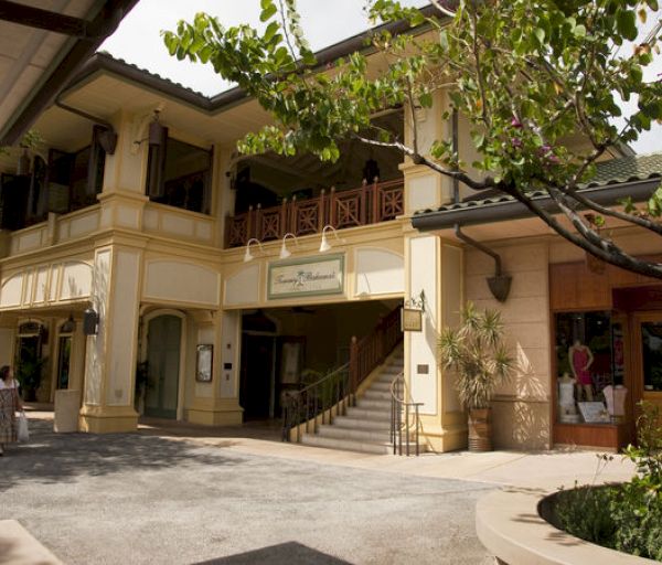 The image shows a two-story yellow building with a staircase, surrounded by greenery and featuring a woman walking on the left side of the image.