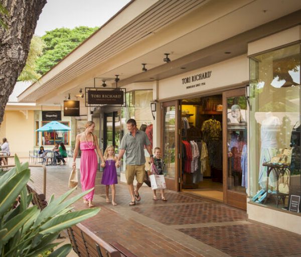 A family is walking near a clothing store in an outdoor shopping center on a sunny day, with some people sitting in the background.