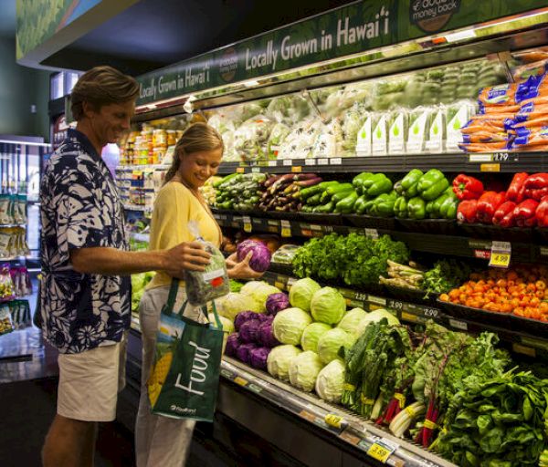 A man and woman are grocery shopping together in the fresh produce section, selecting vegetables and holding a reusable bag.