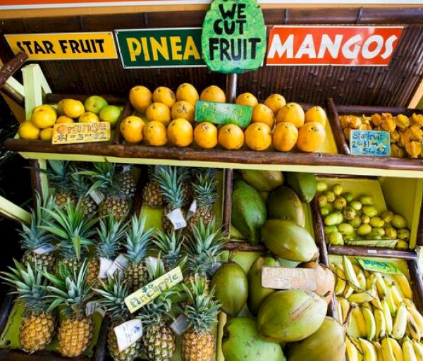 The image shows a fruit stand with star fruits, pineapples, mangoes, oranges, bananas, coconuts, and limes, with a sign saying 