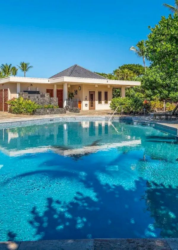 A serene outdoor swimming pool surrounded by lush tropical greenery and lounge chairs, with a small building in the background and clear blue sky.