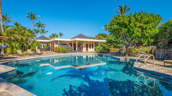 A luxurious outdoor pool with a ladder, surrounded by lush tropical plants, sun loungers, and a modern building is in the background, under a blue sky.