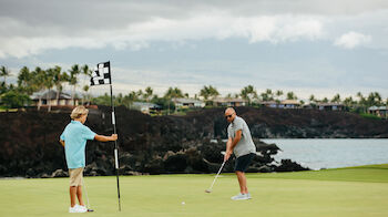 Two people playing golf on a green near a waterfront with trees and houses in the background. The sky appears overcast.