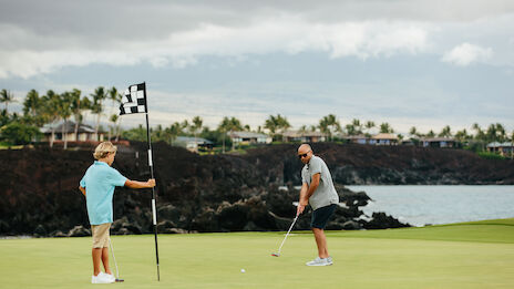 Two people playing golf on a green near a waterfront with trees and houses in the background. The sky appears overcast.