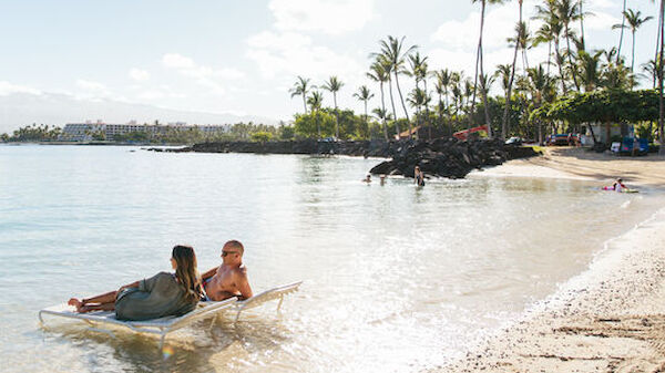 Two people lounge on chairs in shallow, sunlit water near a sandy beach with palm trees and distant buildings under a bright sky.