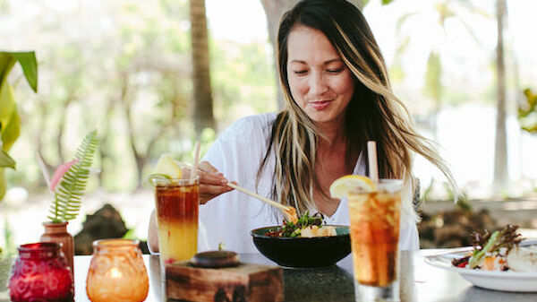 A woman dining outdoors at a beautifully set table. She is enjoying a meal with two different beverages beside her and surrounded by nature.