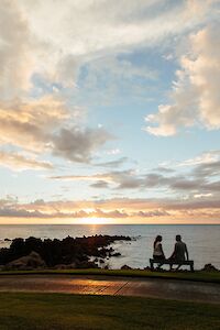 A couple sits on a bench by the ocean, enjoying a sunset with colorful clouds above and rocky shores nearby.