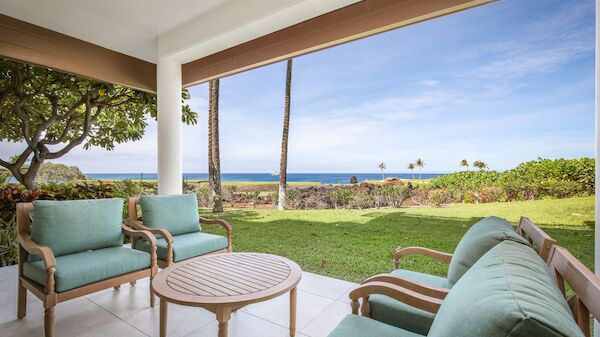 A patio with cushioned chairs and a round table, overlooking green grass, palm trees, and an ocean view under a clear sky.