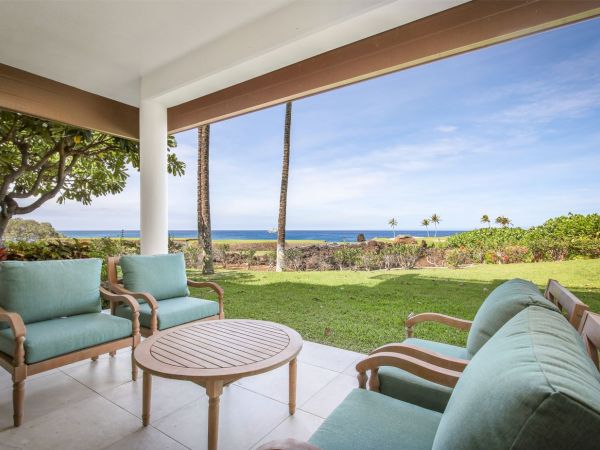 A patio with cushioned chairs and a round table, overlooking green grass, palm trees, and an ocean view under a clear sky.