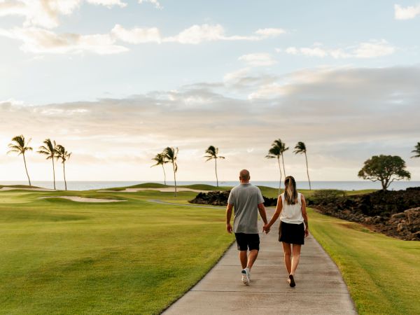 A couple is walking hand in hand on a pathway, surrounded by lush green grass, palm trees, and a scenic ocean view in the background, at sunset.